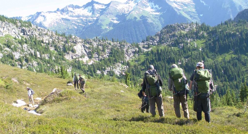 a group of outward bound students wearing backpacks hike along a grassy alpine area. There are vast mountains in the distance.